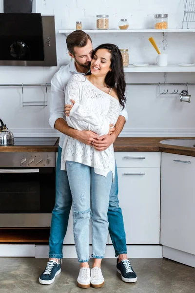 Piena vista lunghezza dell'uomo abbracciando dolcemente donna felice in cucina — Foto stock