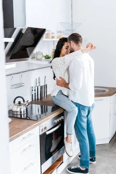 Mujer feliz sentada en la mesa en la cocina y abrazando al hombre - foto de stock