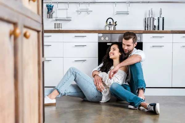 Feliz casal sentado no chão e abraçando na cozinha — Fotografia de Stock