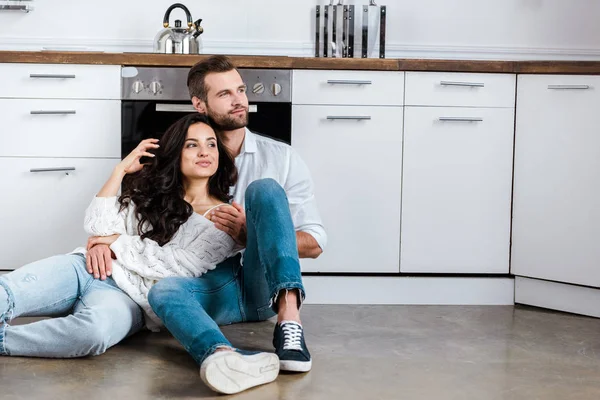 Pareja sentada en el suelo y abrazándose a la cocina y mirando hacia otro lado - foto de stock