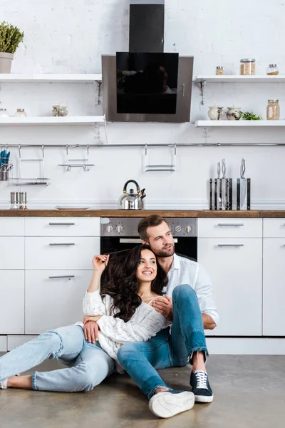 Feliz pareja sentada en el suelo y abrazándose a la cocina y mirando hacia otro lado - foto de stock