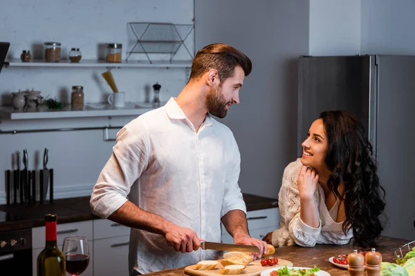 Homem cortando pão na mesa de madeira e olhando para a mulher feliz — Stock Photo