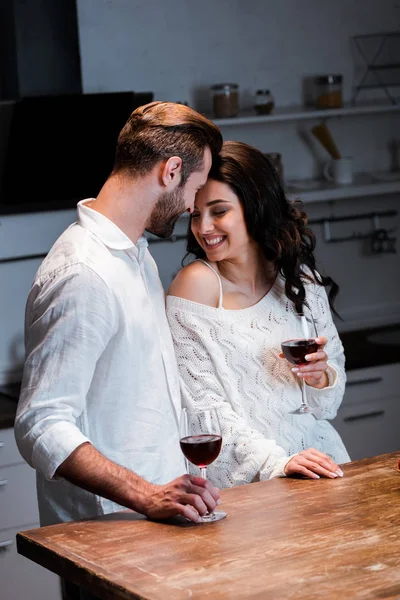 Happy couple holding wineglasses with red wine and gently hugging at kitchen — Stock Photo