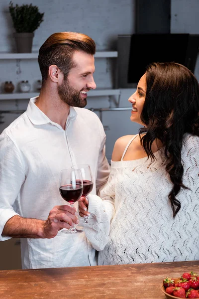 Pareja feliz tintineo con gafas de vino y mirándose unos a otros en la mesa de madera - foto de stock