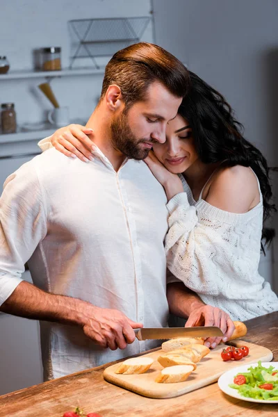 Woman embracing man and looking how boyfriend cutting bread at wooden table — Stock Photo