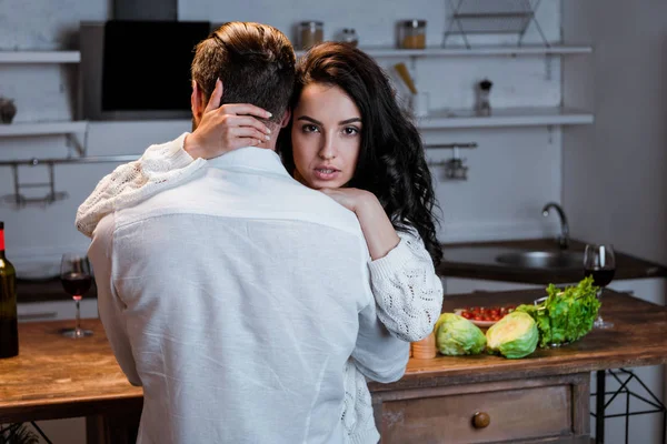 Пристрасна брюнетка жінка обіймає чоловіка і дивиться на камеру на кухні — стокове фото