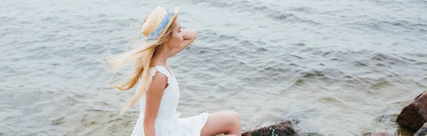 Panoramic shot of dreamy blonde girl touching straw hat while sitting in white dress near sea — Stock Photo