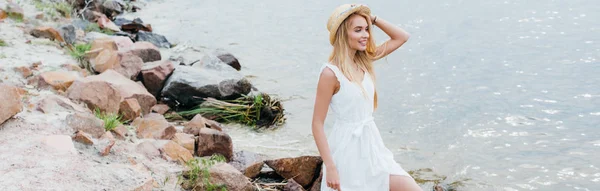 Plano panorámico de alegre joven rubia tocando sombrero de paja y mirando al mar - foto de stock