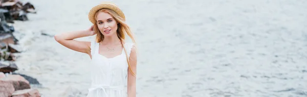 Plan panoramique de joyeux jeune femme blonde touchant chapeau de paille et regardant la caméra près de la mer — Photo de stock