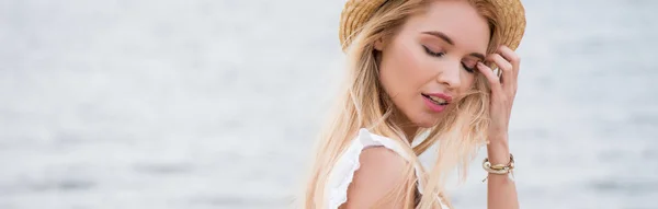 Plan panoramique de jolie femme blonde aux yeux fermés touchant les cheveux près de la mer — Photo de stock