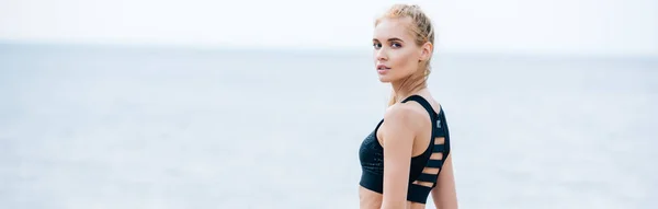 Plan panoramique de jolie jeune femme blonde en vêtements de sport regardant la caméra près de la mer — Photo de stock