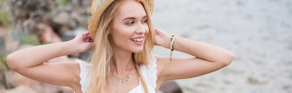 Панорамный снимок счастливой молодой блондинки, трогающей соломенную шляпу — стоковое фото