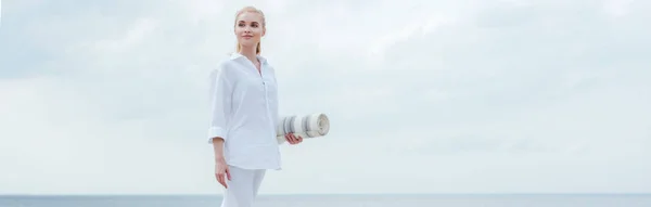 Panoramic shot of happy young blonde woman standing near sea and holding yoga mat — Stock Photo