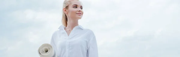 Panoramaaufnahme einer fröhlichen jungen blonden Frau, die am Meer steht und eine Yogamatte hält — Stockfoto