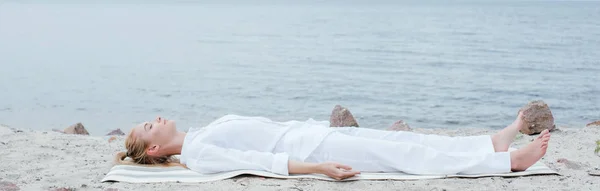 Panoramic shot of attractive blonde woman with closed eyes meditating while lying on yoga mat near sea — Stock Photo