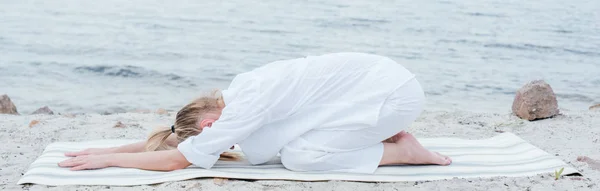 Plano panorámico de mujer joven rubia relajándose y practicando yoga en estera de yoga cerca del mar - foto de stock
