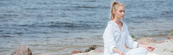 Panoramic shot peaceful blonde woman with closed eyes practicing yoga near river — Stock Photo