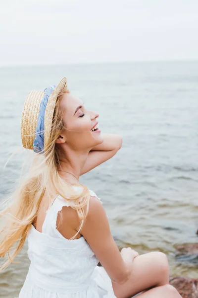 Glückliche junge Frau mit geschlossenen Augen, die lächelt, während sie Strohhut berührt — Stockfoto