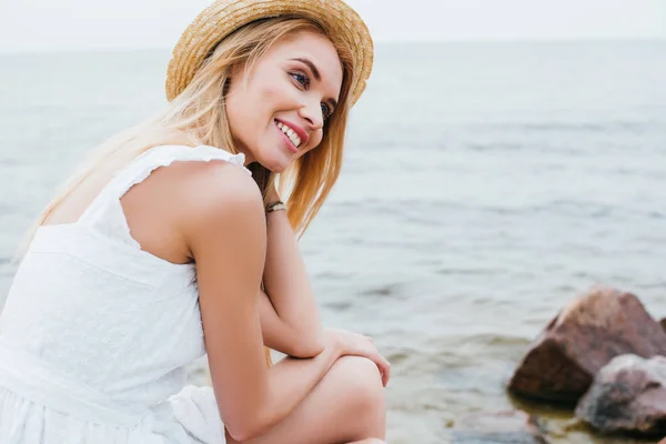 Cheerful blonde woman in white dress and straw hat smiling near sea — Stock Photo