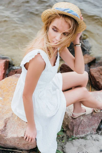 Vue aérienne de jolie fille blonde touchant chapeau de paille tout en étant assis sur des pierres — Photo de stock