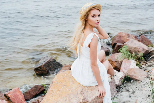 Beautiful blonde girl touching straw hat while sitting on stones near sea — Stock Photo