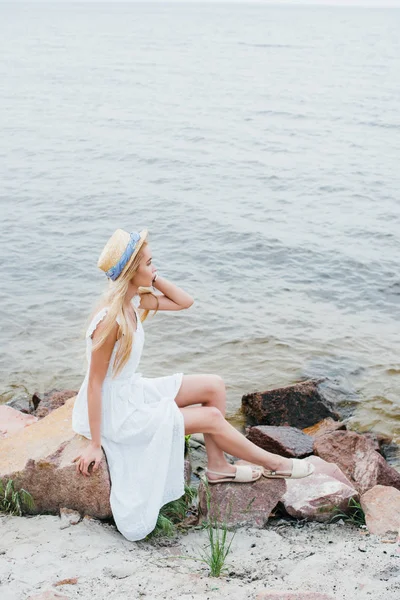 Dreamy blonde girl touching straw hat while sitting on stones near sea — Stock Photo