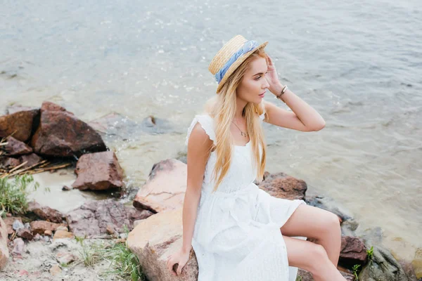 Donna bionda sognante toccare cappello di paglia mentre seduto su pietre vicino al mare — Foto stock