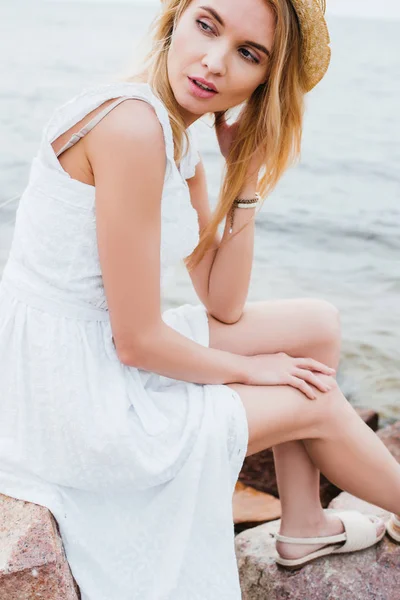 Dreamy woman touching straw hat while sitting on stones near sea — Stock Photo