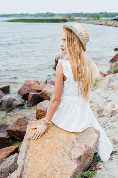 Hermosa mujer rubia en sombrero de paja sentado en piedras cerca del mar - foto de stock