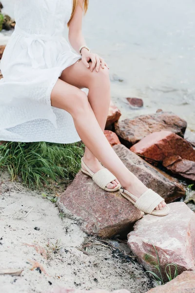 Vista cortada de jovem mulher em vestido branco sentado em pedras perto do mar — Fotografia de Stock