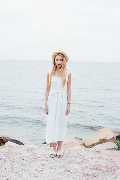 Mujer joven de pie en vestido blanco y sombrero de paja cerca del mar - foto de stock