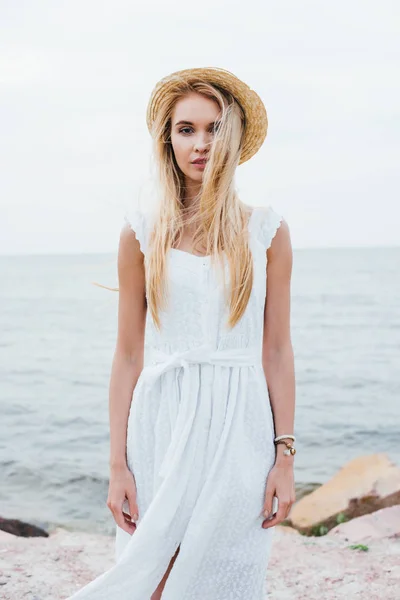 Приваблива молода жінка стоїть в білій сукні і солом'яному капелюсі біля моря — стокове фото