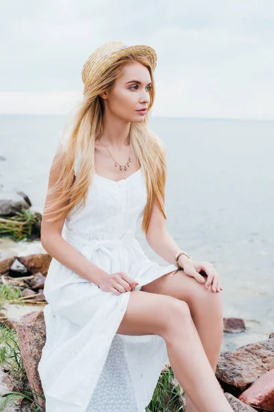 Atractiva mujer rubia en vestido blanco y sombrero de paja sentado cerca del mar - foto de stock