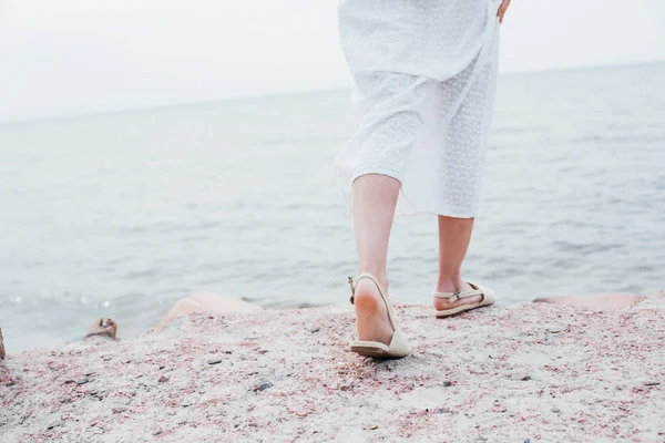 Vista recortada de mujer joven en vestido blanco y sandalias de pie cerca del mar - foto de stock