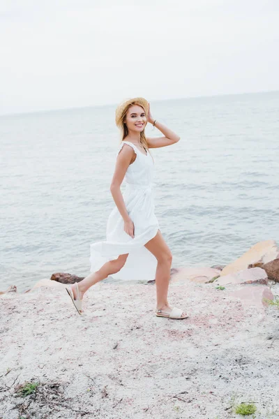 Glückliches blondes Mädchen lächelt, während es Strohhut berührt und am Meer spaziert — Stockfoto