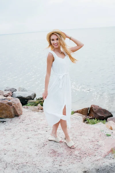 Happy girl smiling while touching straw hat and standing near sea — Stock Photo