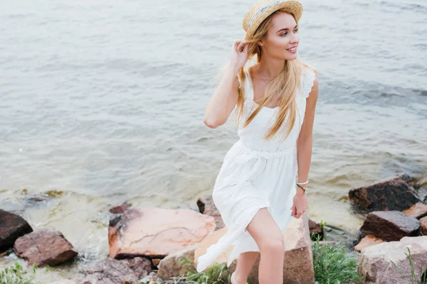 Bonita chica sonriendo mientras toca sombrero de paja y caminando cerca del mar - foto de stock