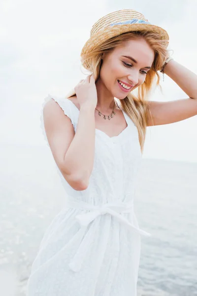 Pretty young woman smiling while touching straw hat near sea — Stock Photo