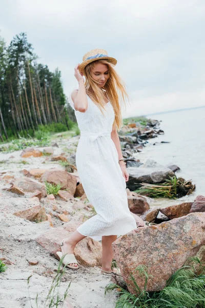 Schöne blonde Frau steht und berührt Strohhut am Meer — Stockfoto