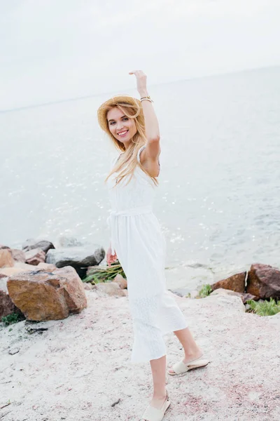 Cheerful blonde woman in straw hat smiling while gesturing near sea — Stock Photo