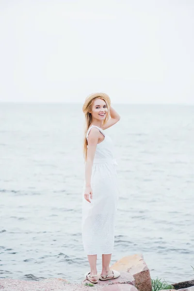 Sonriente rubia joven tocando sombrero de paja y mirando a la cámara mientras está de pie en vestido blanco cerca del mar - foto de stock