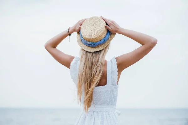 Back view of young blonde woman standing and touching straw hat — Stock Photo