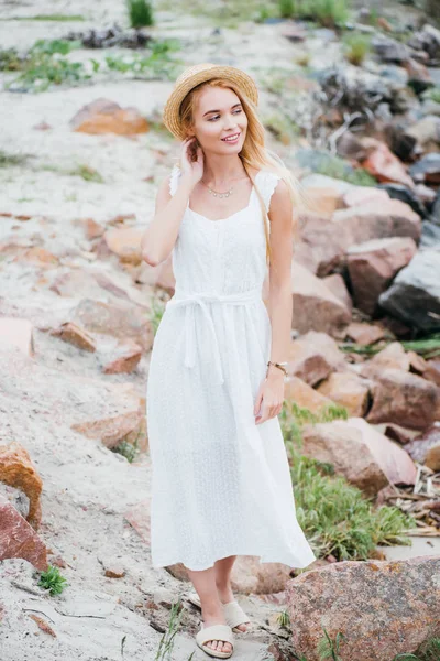 Jovem mulher loira alegre em chapéu de palha em pé sobre pedras em vestido branco — Fotografia de Stock