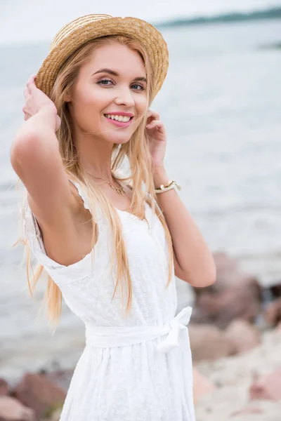 Donna bionda sorridente guardando la fotocamera e toccando cappello di paglia vicino al mare — Foto stock