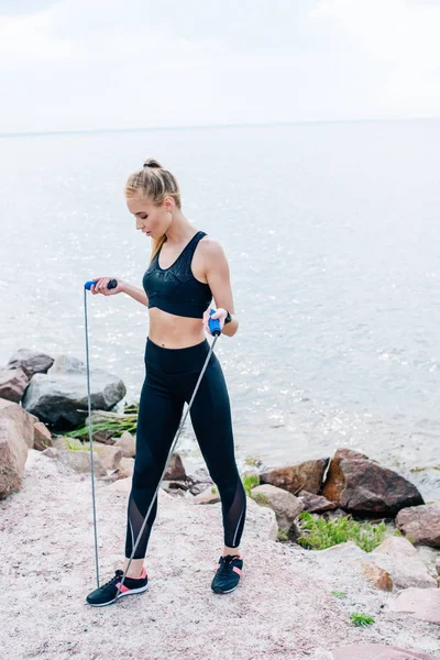 Jolie jeune femme en vêtements de sport debout et tenant corde à sauter près de la mer — Photo de stock