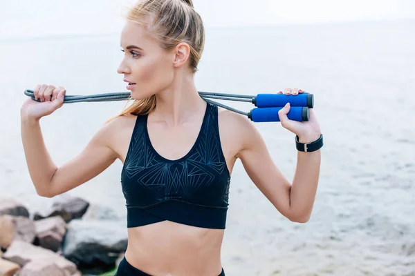 Bonita joven en ropa deportiva sosteniendo saltar la cuerda cerca del mar - foto de stock