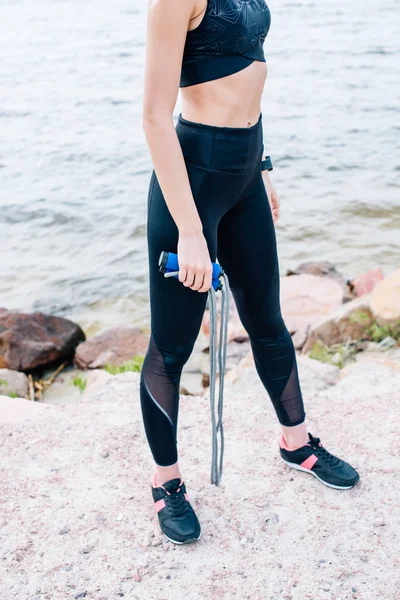 Vista recortada de mujer joven en ropa deportiva sosteniendo saltar la cuerda cerca del mar - foto de stock