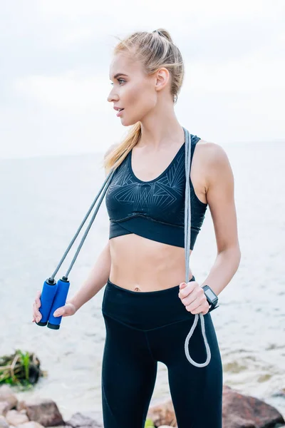 Hermosa joven en ropa deportiva sosteniendo saltar la cuerda cerca del mar - foto de stock