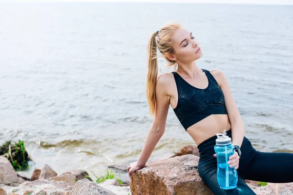 Blonde sportswoman holding sport bottle while sitting on stone near sea — Stock Photo