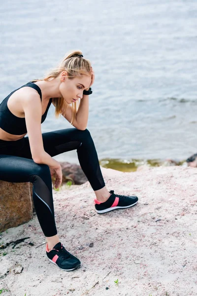 Exhausted blonde young sportswoman sitting on stone near sea — Stock Photo